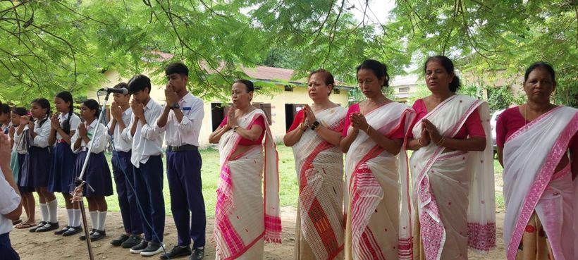 Teachers in white mekhela chador and students in uniform with hands folded in prayer under the trees